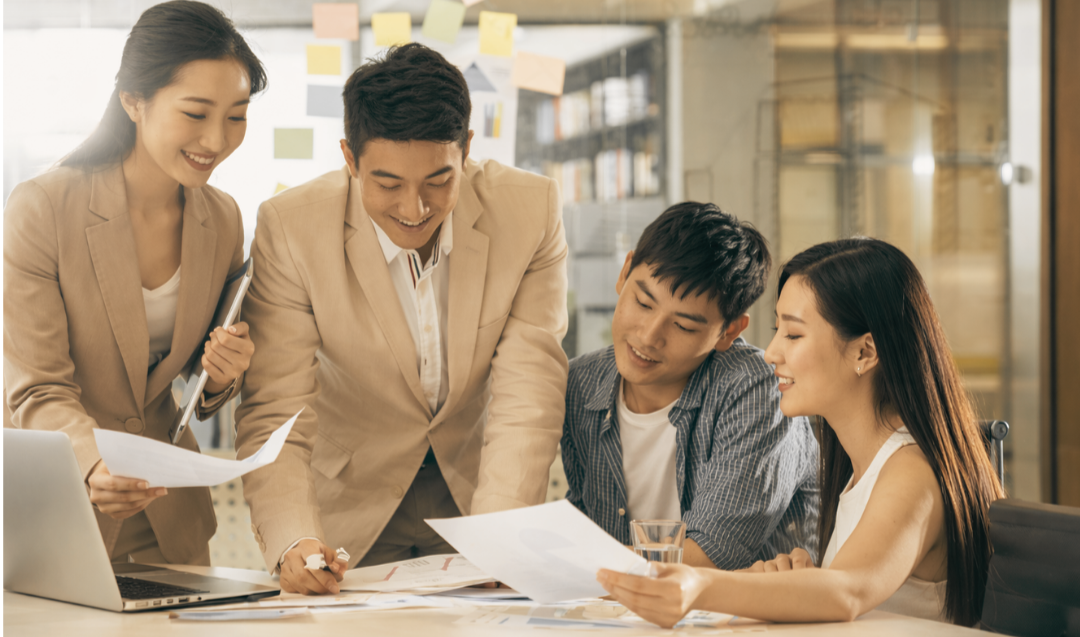 Group of four people in a modern office setting, discussing documents around a laptop.